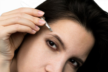 Beautiful young brunette and dark-eyed woman plucking her eyebrows with steel tweezers. Personal care. Suffering during depilation. Modern standards of beauty.