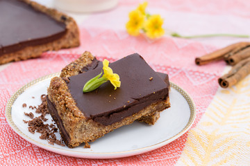 Сhocolate tart made from oatmeal, coconut milk and butter, dates and almonds on a white saucer.Cinnamon sticks and yellow flowers in the background.Сoncept of healthy food and healthy dessert 
