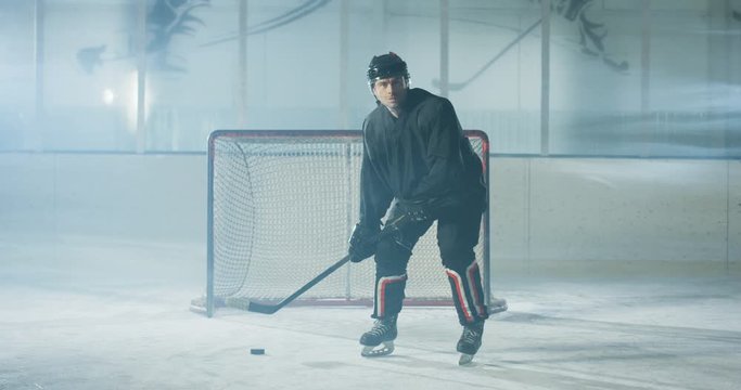Portrait of Caucasian handsome concentrated male hockey player in uniform and helmet standing on ice arena with club and puck. Sportsman in casque with stick and disc on icy surface. Goalkeeper.