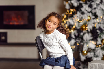 Beautiful little girl standing at the large fireplace and holding a gift, in the bright New Year's interior