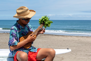 Japanese Teenage boy playing the Ukulele on the beach in Hebara, Chiba, Japan. There is a white surfboard & perfect blue ocean in the background, he has a sun tan & nice smile.