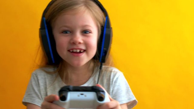 little blonde with blue eyes in headphones plays a video game with a gamepad on a yellow background.