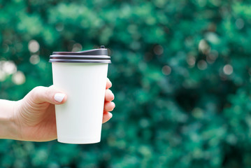 Female hand holds paper cup of coffee on a blurred green background.