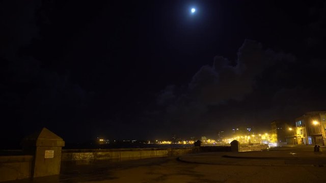 Havana Cuba time lapse along the Malecon waterfront at night, cars driving by, people hanging out, some apartment buildings in the background and a bright moon in the sky.