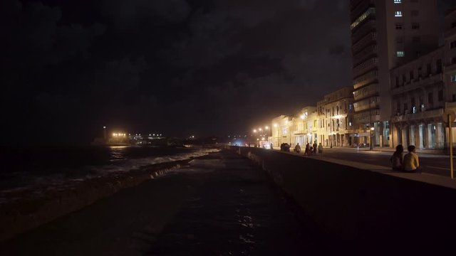 Havana Cuba time lapse along the Malecon waterfront at night, cars driving by, people hanging out, some apartment buildings in the background and Morro Castle and lighthouse off in the distance.