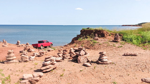 A red pick up truck is parked on the red sand beach of North Cape, a destination just north of Tignish, PEI.  The truck is surrounded by the mini inukshuk statues that are scattered about the beach.