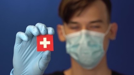 COVID-19 pandemic in Switzerland. A young, attractive Swiss man in protective medical mask and gloves holding the flag of Swiss Confederation. Coronavirus pandemic in Europe - Powered by Adobe