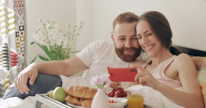 Happy guy and girl watching funny video and laughing in bed. Young romantic family having breakfast and talking while looking at smartphone screen in early morning. Concept of leisure.