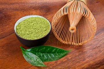 Powdered matcha tea in a cup and green leaves with bamboo matcha tea whisk also know as chasen on a wooden background.
