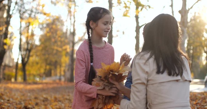 Caucasian brunette mother in white trench and her two daughters sit in city park. Girls collect fallen leaves and give bouquets to mom. Carpet of leaves. Positive emotions 50 fps slow motion