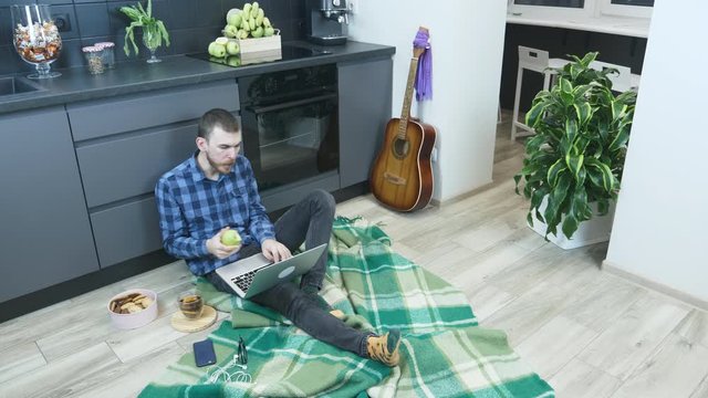 Young bearded man using laptop during work on remote project at home. Male with computer, working on distance sitting on floor in kitchen, eating apple and having fun. Working at home at quarantine