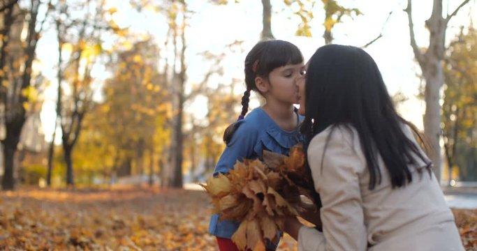 Caucasian brunette mother in white trench and her daughter in city park. Girl collecting fallen leaves and give bouquet to mom. Carpet of leaves. Positive emotions 50 fps slow motion