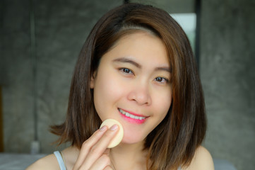 Portrait of young attractive Asian woman applying foundation powder puff on her face to cover flaws and change the natural skintone. Conceptual of women's beauty and cosmetic.