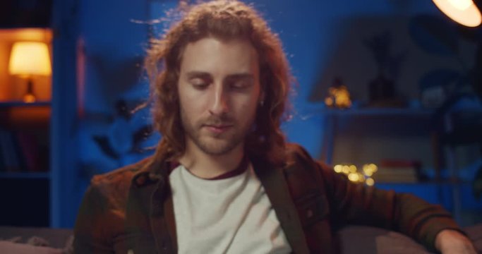 Portrait of long haired hipster guy looking at smartphone screen and raising head. Handsome young man smiling to camera while holding moile phone and sitting on couch at home. Zoom in.