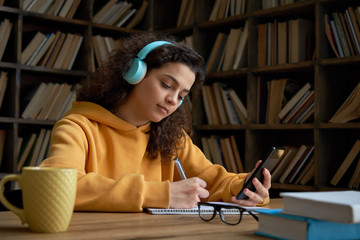 Latin teen girl wear headphones hold phone online learning in mobile app. Hispanic college student...