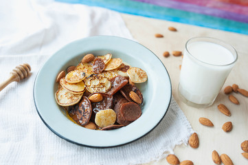 tiny pancakes with almond nuts and honey in metal plate on bright kitchen table with milk cup, scattered nuts, white napkin and honey stick. trending food cereal pancakes.