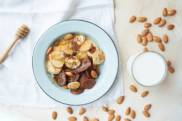 tiny pancakes with almond nuts and honey in metal plate on bright kitchen table with milk cup, scattered nuts, white napkin and honey stick. trending food cereal pancakes. top view, flat lay