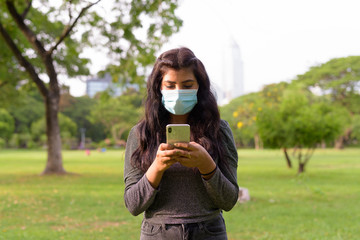 Young Indian woman with mask using phone while relaxing at the park