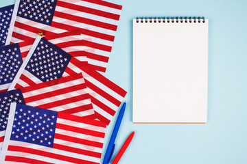 Top view on set of american flags, red and blue pens and empty notebook with copyspace for text on light blue background. National symbol of USA - flag Old Glory. Business or education concept