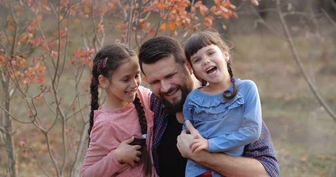 Portrait of father with two daughters in beautiful autumn forest. Bearded man in plaid shirt  hugging little girls, smiling, having fun looking to camera. Father's day concept. 50 fps slow motion 4k