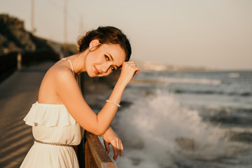 Portrait of beautiful young smiling girl in the white dress in greek style walking along the sea promenade. Summer, sunset time. Bride, wedding at sea 