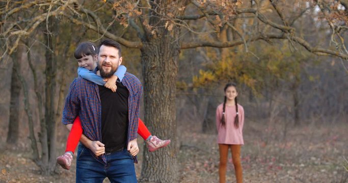 Father with two daughters walking in beautiful autumn forest. Bearded man riding a girl of preschool age on his back and playing a horse . Father's day concept. 50 fps slow motion 4k
