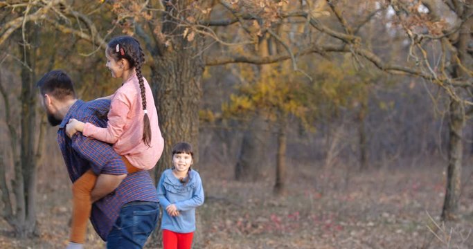 Father with two daughters walking in beautiful autumn forest. Bearded man riding a girl of preschool age on his back and playing a horse . Father's day concept. 50 fps slow motion 4k