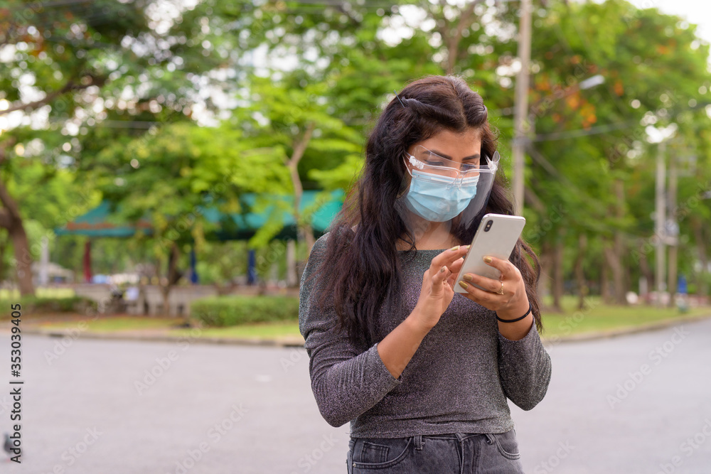 Wall mural Young Indian woman with mask and face shield using phone at the park outdoors