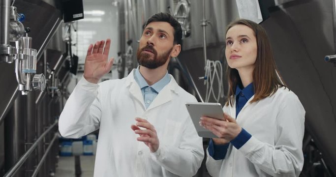 Brewery workers in white lab coats checking equipment. Man talking and giving instructions and woman using tablet while standing at rows of steel brewing vats and looking aside