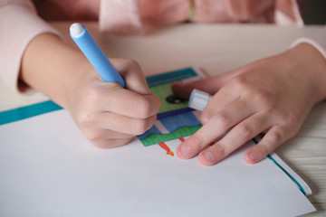A girl in a pink sweatshirt draws with a light blue felt-tip pen at a table. Blurred front background. Beige table