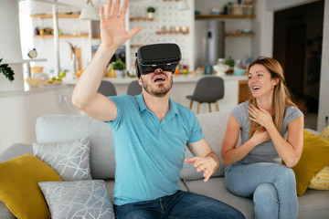 Obraz premium Smiling young man using VR headset at home on couch. Man and his wife enjoying virtual reality at her apartment. 