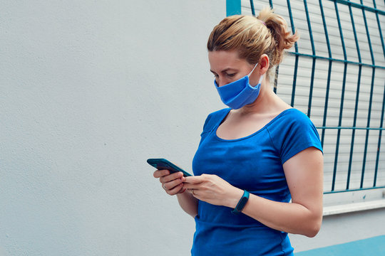 blonde white woman in a surgical mask walking down the street looking at her cell phone