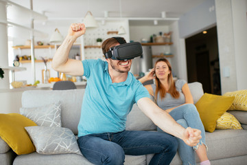 Smiling young man using VR headset at home on couch. Man and his wife enjoying virtual reality at her apartment.	
