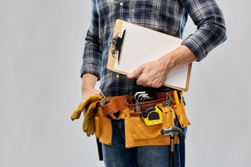 repair, construction and building - male worker or builder with clipboard and working tools on belt over grey background