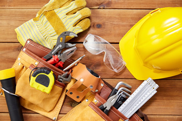 repair, building and renovation concept - gloves, protective glasses, helmet and belt with different work tools in pockets on wooden boards background