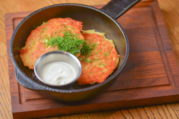 Delicious potato pancakes with sour cream served in a frying pan on wooden board over light rustic wooden table.