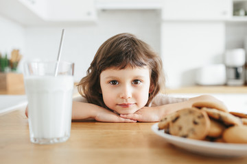 Cute toddler girl drinking milk with steel straw from glass and eating cookies