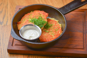 Delicious potato pancakes with sour cream served in a frying pan on wooden board over light rustic wooden table.