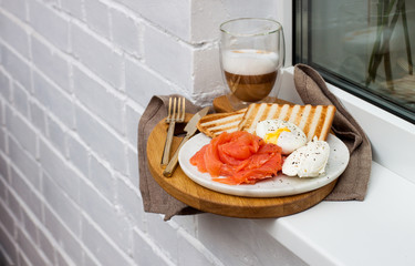 Toasted bread, salted salmon and poached egg with cream cheese and cup of coffee on white windowsill.