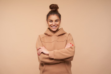 Fototapeta premium Good looking young happy brown haired lady showing her white perfect teeth while smiling cheerfully and crossing hands on her chest, posing over beige background