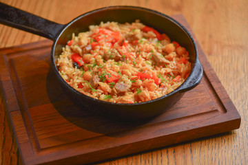 Rice with meat and vegetables served in a frying pan on wooden board over light rustic wooden table