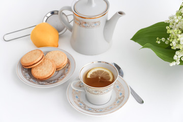 Porcelain tea set with a cup, teapot on white tablecloth with cookies on a plate and lilies of the valley on table. Afternoon tea party concept. Breakfast. Morning. Home sweet home.