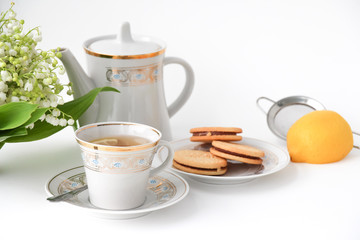 Porcelain tea set with a cup, teapot on white tablecloth with cookies on a plate and lilies of the valley on table. Afternoon tea party concept. Breakfast. Morning. Home sweet home.