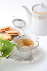 Porcelain tea set with a cup, teapot on white tablecloth with cookies on a plate and lilies of the valley on table. Afternoon tea party concept. Breakfast. Morning. Home sweet home.