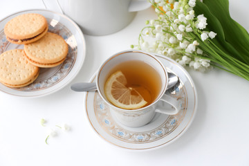 Porcelain tea set with a cup, teapot on white tablecloth with cookies on a plate and lilies of the...