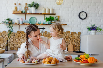Family portrait of a happy mother and daughter posing in the kitchen during Breakfast, eating delicious macaroons, cakes, cookies.