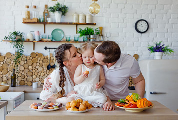 Family portrait of a happy mother, daughter and father posing in the kitchen during Breakfast, eating delicious macaroons, cakes, cookies. Married good relations, love each other