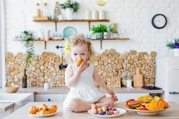 A little girl poses in the kitchen during Breakfast, eating delicious macaroons, cakes, cookies.