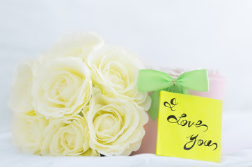 Romantic present. Bouquet of white roses, gift box and "I Love You" note placed on a bed. Closeup.