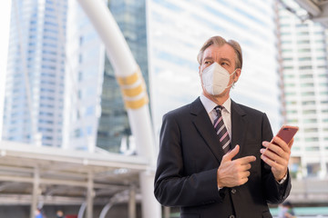 Mature businessman with mask thinking while using phone at skywalk bridge